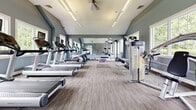 Located at 9 Royal Court in Wayne, NJ, this well-appointed fitness center offers a comprehensive workout environment within a single-floor layout designed for both functionality and comfort. The space is characterized by striking vaulted ceilings that lend an open, airy feel, complemented by soft blue-gray walls and neutral-toned carpet flooring that runs throughout.

The cardio zone is prominently featured along one side of the room, housing a row of treadmills, an elliptical, and a stationary bike, all facing a full wall of floor-to-ceiling mirrors that visually expand the space. The mirrors also provide excellent form-checking capability for users during workouts.

The strength training area is equally well-equipped, featuring a cable machine, weight bench, and a varied collection of kettlebells and free weights neatly arranged on the floor. Dedicated exercise mats provide space for stretching and bodyweight training.

Natural light fills the facility through multiple windows and a glass-panel entry door that opens to an outdoor view, contributing to an inviting and energizing atmosphere. A ceiling fan aids in ventilation, and recessed and linear lighting ensures the space is well-lit at all times. A water fountain near the entrance adds a practical amenity. The overall design is clean, modern, and purpose-built to support a full range of fitness activities.