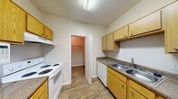 This single-floor residence offers a well-organized and functional layout designed for comfortable everyday living. The kitchen is a standout area, featuring warm honey-toned wood cabinetry that lines both walls, dark granite-style countertops, and wood-look flooring throughout. White appliances — including a coil-top range, dishwasher, refrigerator, and built-in microwave — provide a complete cooking setup. A double-basin sink sits beneath upper cabinets, and a dedicated walk-in pantry or storage closet adds convenient organization space. The kitchen opens toward the main living area, which is carpeted and brightened by natural light streaming through a window fitted with blinds. A ceiling fan overhead adds comfort and airflow. The bedrooms are carpeted and neutral in tone, each offering a calm and flexible space. One bedroom features a sliding-door closet with a rod and shelf, while another benefits from a window with views of surrounding greenery. The bathroom is well-appointed with a dual-sink vanity topped with matching granite-style counters, warm wood cabinetry, a large mirror, wood-look flooring, and a separate shower/tub area. Throughout the unit, a consistent neutral color palette and cohesive finishes create a clean, move-in-ready atmosphere.