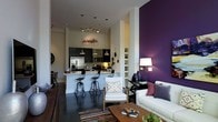 This open-concept space makes a strong visual statement through thoughtful design choices and a cohesive color palette. The living area is anchored by a dramatic deep-purple accent wall that creates a rich backdrop for the seating arrangement, which includes a cushioned sofa adorned with decorative pillows and a clean-lined armchair positioned around a central coffee table. A floor lamp provides warm ambient lighting in the lounge area, complementing the recessed ceiling lights throughout.

The kitchen flows seamlessly from the living space, featuring dark cabinetry, stainless steel appliances including a refrigerator and microwave, and light countertops. A raised breakfast bar with stylish black metal counter stools serves as both a functional dining area and a natural divider between the kitchen and living zones.

The neutral white walls and light ceiling contrast effectively with the darker accents throughout, maintaining brightness across the space. A colorful area rug grounds the living area, adding warmth and texture to the dark flooring. Open shelving along one wall provides display and storage opportunities, while pendant lighting above the kitchen bar adds refined detail to the overall design.