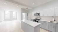 Located on Broadway in Troy, NY, this single-floor unit offers a thoughtfully designed layout with modern finishes throughout. The open-concept kitchen serves as the central feature of the space, showcasing two-tone cabinetry — white upper cabinets paired with gray lower cabinets — topped with crisp white countertops. A center island provides additional workspace and acts as a natural divider between the kitchen and living areas. Stainless steel appliances, including a range, over-the-range microwave, and refrigerator, complete the kitchen. A matte black faucet adds a contemporary accent above the undermount sink. Light-toned luxury vinyl plank flooring runs throughout the unit, contributing to a bright, cohesive aesthetic. Recessed lighting illuminates the kitchen and living areas, while large windows draw in natural light from the Broadway-facing side of the unit. Additional rooms offer clean, neutral spaces with soft gray walls and white trim. The bathroom features a walk-in shower dressed in large-format marble-look tile with a classic white hex mosaic floor, conveying a polished, upscale feel. The overall ambiance is clean, modern, and move-in ready.