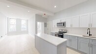 Located on Broadway in Troy, NY, this single-floor unit offers a thoughtfully designed layout with modern finishes throughout. The open-concept kitchen serves as the central feature of the space, showcasing two-tone cabinetry — white upper cabinets paired with gray lower cabinets — topped with crisp white countertops. A center island provides additional workspace and acts as a natural divider between the kitchen and living areas. Stainless steel appliances, including a range, over-the-range microwave, and refrigerator, complete the kitchen. A matte black faucet adds a contemporary accent above the undermount sink. Light-toned luxury vinyl plank flooring runs throughout the unit, contributing to a bright, cohesive aesthetic. Recessed lighting illuminates the kitchen and living areas, while large windows draw in natural light from the Broadway-facing side of the unit. Additional rooms offer clean, neutral spaces with soft gray walls and white trim. The bathroom features a walk-in shower dressed in large-format marble-look tile with a classic white hex mosaic floor, conveying a polished, upscale feel. The overall ambiance is clean, modern, and move-in ready.