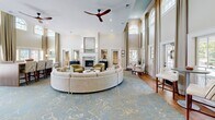 Located at Granby Crossing in Cayce, South Carolina, this beautifully appointed single-floor space makes a grand impression with its dramatic double-height ceilings and sweeping arched transom windows that flood the interior with natural light. The expansive living room is the centerpiece of the space, showcasing floor-to-ceiling draped windows, warm hardwood flooring, and a refined color palette of soft neutrals accented by subtle gold and blue tones. A curved sectional sofa anchors the seating area, complemented by plush accent chairs in sage green and a decorative area rug. A marble-surround fireplace adds an elegant focal point, while a ceiling fan overhead provides both function and style. French doors open to views of an outdoor pool area, seamlessly connecting the interior to the exterior. A high-top communal table with upholstered seating offers a more structured gathering space within the room. The kitchen features sage green shaker-style cabinetry with gold hardware, mirrored tile backsplash, open floating wood shelves, and warm wood-paneled ceiling detail. Stainless steel appliances and a stainless under-counter refrigerator complete the well-equipped kitchenette. Together, these spaces offer a welcoming, sophisticated environment ideal for community gathering and relaxation.