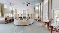 Located at Granby Crossing in Cayce, South Carolina, this beautifully appointed single-floor space makes a grand impression with its dramatic double-height ceilings and sweeping arched transom windows that flood the interior with natural light. The expansive living room is the centerpiece of the space, showcasing floor-to-ceiling draped windows, warm hardwood flooring, and a refined color palette of soft neutrals accented by subtle gold and blue tones. A curved sectional sofa anchors the seating area, complemented by plush accent chairs in sage green and a decorative area rug. A marble-surround fireplace adds an elegant focal point, while a ceiling fan overhead provides both function and style. French doors open to views of an outdoor pool area, seamlessly connecting the interior to the exterior. A high-top communal table with upholstered seating offers a more structured gathering space within the room. The kitchen features sage green shaker-style cabinetry with gold hardware, mirrored tile backsplash, open floating wood shelves, and warm wood-paneled ceiling detail. Stainless steel appliances and a stainless under-counter refrigerator complete the well-equipped kitchenette. Together, these spaces offer a welcoming, sophisticated environment ideal for community gathering and relaxation.