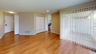 This single-floor residence offers a well-organized layout with consistent design elements throughout. Warm hardwood-style flooring flows seamlessly across the living areas and bedrooms, contributing to a bright, cohesive aesthetic. Neutral beige tones on the walls complement the light flooring, creating an open and inviting atmosphere.

The kitchen is a standout feature, equipped with white shaker-style cabinetry, granite countertops with subtle speckled detailing, and a full suite of stainless steel GE appliances, including a range, over-the-range microwave, and refrigerator. The layout is efficiently designed to maximize counter and storage space.

The living area is generously proportioned, featuring a set of vertical blinds that lead to what appears to be a patio or exterior access point, along with closet storage. Adjacent rooms, likely bedrooms, are well-sized with large windows that allow natural light and views of surrounding greenery.

The bathroom includes a full-size soaking tub with an overhead shower, white tile surround, and chrome fixtures, maintaining a clean and functional design. Overall, the space presents a tidy, move-in-ready environment with practical finishes throughout.