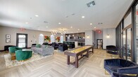 This single-floor club room in Metairie, Louisiana, is a thoughtfully designed social and recreation space that blends modern aesthetics with comfortable functionality. The open-concept layout is anchored by light-toned herringbone hardwood-style flooring that flows throughout, lending warmth and visual continuity to the entire space.

The lounge area features multiple seating arrangements, including a large gray sectional sofa, plush navy armchairs with gold-accented legs, and emerald green velvet barrel chairs arranged around a gold coffee table. Vibrant abstract artwork adorns the walls, adding bold pops of color. Arched windows along one wall flood the space with natural light while offering a classic architectural detail that complements the contemporary interior.

A standout wet bar area is finished with dark charcoal cabinetry, brass hardware, a white quartz countertop, a deep utility sink, and built-in beverage refrigerators. The dramatic dark hexagonal tile backsplash, wall-mounted TV, and brass sconce lighting make this a stylish focal point. A long communal island with pendant lighting provides additional gathering space nearby.

A shuffleboard table occupies a central position in the room, while a glass-walled fitness center is visible at the rear, adding a functional amenity to this well-appointed community gathering space.