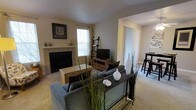This single-floor residence centers around a well-appointed living room with neutral-toned walls and carpeted flooring that creates a warm, inviting atmosphere. A tile-surround fireplace serves as the room's focal point, flanked by a window that allows natural light to filter in. The living area is generously sized, accommodating a sectional sofa arrangement alongside a woven coffee table and a sleek console table with decorative accessories. A lean ladder shelf and media console with a mounted flat-screen television provide functional storage and entertainment options. Tall ornamental grasses and ceramic vases add natural, decorative touches throughout the space. An open passageway connects the living room to an adjacent dining area, visible in the background and furnished with a bar-height table and stools, contributing to the open-concept feel. Vertical blinds on a sliding glass door in the adjoining space suggest access to an outdoor area and provide additional natural light. The cohesive design, neutral palette, and thoughtful layout make this a functional and comfortable living environment suited for everyday living and entertaining.