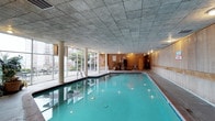 This indoor pool facility offers a well-appointed aquatic environment designed for both recreation and lap swimming. The centerpiece is a generously sized rectangular pool with crystal-clear turquoise water, illuminated by a series of underwater lights that cast a warm, inviting glow throughout the space.

Large floor-to-ceiling windows line one side of the room, flooding the interior with abundant natural light and providing views of the surrounding outdoor area. Sturdy white columns run along the windowed wall, adding an architectural rhythm to the space. The opposite side of the pool is finished with warm wood paneling, lending a classic, refined character to the room.

A textured tile ceiling spans the entire space, contributing to the room's acoustic and moisture management qualities. Pool entry is made convenient with chrome handrails and built-in steps. Along the wood-paneled wall, a bulletin board, potted greenery, and what appears to be a sauna or utility room door provide functional amenities for facility users.

The overall ambiance is clean, bright, and purposeful — a well-maintained aquatic space that balances functionality with a comfortable, welcoming atmosphere suited for regular swimming and leisure use.