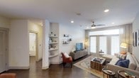 This single-floor space showcases a thoughtfully designed open living area that blends functionality with modern aesthetics. The living room is anchored by rich, dark hardwood flooring that extends throughout, providing a warm contrast to the bright white walls and ceiling. A ceiling fan with a brushed nickel finish adds both style and practicality overhead, while recessed lighting ensures the space is well-lit at all times.

A standout architectural feature is the built-in shelving unit integrated into a structural column, offering tiered display space for décor and personal items. This detail adds character and maximizes vertical space without encroaching on the floor plan. Large windows dressed with horizontal blinds allow generous natural light to fill the room, creating an airy, welcoming atmosphere.

The living area comfortably accommodates a seating arrangement, media console, and a patterned area rug that adds a Southwestern touch. A glimpse through an open doorway reveals a bathroom with a soaking tub, indicating a well-appointed layout. Overall, the space presents a clean, contemporary feel with warm, inviting design elements throughout.