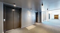 This common area hallway at 2125 Rue Ward in Montréal, QC, is a well-designed corridor that serves as a functional and visually appealing shared space within the building. The hallway is defined by a striking contrast between two distinct zones: an interior corridor and a light-filled glazed passage running alongside it.

The interior section features smooth grey walls, dark ceiling panels with recessed and surface-mounted spotlights, and warm-toned carpet flooring. Elevator doors finished in dark metal provide access between floors, and informational panels are mounted neatly on the walls, contributing to a clean, organized aesthetic.

Transitioning toward the glazed section, the hallway opens dramatically through a continuous floor-to-ceiling curtain wall of large-pane windows supported by exposed concrete columns. This wall of glass floods the space with abundant natural light and offers clear views of the surrounding neighbourhood and adjacent building facades with balconies. The blue-toned striped carpet in this section complements the bright, airy atmosphere created by the expansive glazing.

Together, the two zones create a cohesive yet dynamic circulation space that balances privacy and openness. The combination of modern materials — concrete, glass, carpet, and metal — reflects a contemporary architectural approach suited to a well-appointed residential building.
