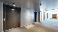 This common area hallway at 2125 Rue Ward in Montréal, QC, is a well-designed corridor that serves as a functional and visually appealing shared space within the building. The hallway is defined by a striking contrast between two distinct zones: an interior corridor and a light-filled glazed passage running alongside it.

The interior section features smooth grey walls, dark ceiling panels with recessed and surface-mounted spotlights, and warm-toned carpet flooring. Elevator doors finished in dark metal provide access between floors, and informational panels are mounted neatly on the walls, contributing to a clean, organized aesthetic.

Transitioning toward the glazed section, the hallway opens dramatically through a continuous floor-to-ceiling curtain wall of large-pane windows supported by exposed concrete columns. This wall of glass floods the space with abundant natural light and offers clear views of the surrounding neighbourhood and adjacent building facades with balconies. The blue-toned striped carpet in this section complements the bright, airy atmosphere created by the expansive glazing.

Together, the two zones create a cohesive yet dynamic circulation space that balances privacy and openness. The combination of modern materials — concrete, glass, carpet, and metal — reflects a contemporary architectural approach suited to a well-appointed residential building.
