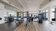 Located at 327–349 W Tremont Ave in Charlotte's South End neighborhood, this fitness center offers a well-appointed, single-floor facility designed to support a wide range of workout routines. The main exercise floor is expansive and bright, featuring soaring ceilings with exposed white ductwork and pendant lighting that lend an industrial-chic aesthetic. Large windows line the perimeter, flooding the space with natural light and providing views of the surrounding streetscape.

The cardio and strength training area is outfitted with treadmills, ellipticals, cable machines, weight benches, and power racks with plate-loaded barbells, all arranged on durable hardwood-style flooring. Full-length mirrors amplify the sense of space and support proper form during workouts. A dedicated functional training zone, framed by warm wood slat partitions, houses a rack stocked with medicine balls, kettlebells, battle ropes, and resistance bands, along with open floor space for dynamic training and stretching.

A separate, more intimate studio area features rubber flooring and a wall-mounted television, ideal for group classes or personal training sessions. The facility also includes a built-in hydration station with a stainless steel double water fountain and bottle filler, as well as storage cabinetry and motivational sports photography displayed along the walls, contributing to an energetic and polished atmosphere throughout.