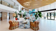 Located at 4341 34th St S in Saint Petersburg, Florida, this sophisticated two-story commercial space offers an expansive and elegantly designed interior spread across two floors. The ground floor commands attention with a soaring double-height atrium that floods the space with natural light through large glass entry doors and a striking multicolored stained-glass feature wall, which serves as a vivid focal point visible from both levels. A sweeping staircase with glass railings and metal balustrades connects the floors, while elevator access provides additional convenience.

The main lobby is anchored by a sculptural slatted wood ceiling installation that curves organically overhead, complemented by contemporary ring chandeliers. Lush tropical plants, curved white sofas, rattan accent chairs, and layered area rugs create a warm, resort-inspired atmosphere. Large-format porcelain tile flooring extends throughout, maintaining a clean and cohesive aesthetic.

Vertical slatted wood wall panels line corridors and accent walls on both levels, introducing warmth and texture against the crisp white structural columns and ceilings. The upper floor features an open mezzanine corridor with glass railings overlooking the grand lobby below, as well as additional hallway and flex spaces. The overall design balances modern architectural precision with coastal Florida warmth.