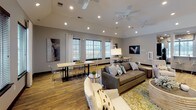 This single-floor community space offers a welcoming and well-appointed environment designed for both relaxation and social interaction. The open-concept main lounge is generously proportioned, featuring soaring ceilings equipped with multiple ceiling fans and recessed lighting that ensure a bright, airy atmosphere. Rich hardwood-style flooring runs throughout, complementing the warm, neutral gray walls and contemporary décor.

The seating area is anchored by a plush gray sofa and accent chairs arranged around a central coffee table, while a striking stacked stone accent wall serves as a bold architectural focal point. Multiple dining tables with modern acrylic and metal chairs provide ample space for gatherings. Large windows dressed with grommet-top curtains and horizontal blinds line the perimeter, flooding the room with natural light and offering outdoor views.

A dedicated kitchen area features dark espresso cabinetry paired with light granite countertops, stainless steel appliances including a refrigerator, wall oven, and microwave, and durable hardwood-style flooring. A sizable window above the counter further enhances natural light in the space.

Glass-paneled entry doors add a refined, transparent touch to transitions between zones, while carefully curated artwork and decorative accents throughout contribute to the space's polished, modern aesthetic.