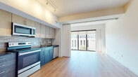 This contemporary single-floor unit offers a well-designed open-concept layout that seamlessly connects the kitchen and living areas. Light hardwood flooring runs throughout, lending warmth and continuity to the space. The living area is bright and airy, anchored by expansive floor-to-ceiling windows fitted with roller shades that invite abundant natural light and provide views of the surrounding streetscape. A sliding panel partition offers flexible separation between the kitchen and living zone.

The kitchen is efficiently laid out and thoughtfully appointed, featuring two-tone cabinetry — warm upper cabinets paired with dark lower cabinets — complemented by a tile backsplash and dark countertops. Stainless steel appliances, including a refrigerator, electric range, over-the-range microwave, and dishwasher, complete the culinary space. Track lighting overhead adds both function and a modern aesthetic touch.

The bathroom presents a striking design contrast, showcasing a bold black marble-veined countertop set atop natural wood cabinetry. An illuminated frameless mirror enhances the clean, modern finish of the space. Throughout the unit, the clean white walls, contemporary fixtures, and cohesive material palette create a polished and inviting ambiance well-suited for modern urban living.