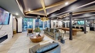 This 1,230-square-foot single-floor amenity lounge at 1200 Riverfront Dr in Little Rock, Arkansas, delivers a sophisticated, multi-functional communal space defined by high-end architectural detailing and thoughtful zoning. The ceiling is a standout feature throughout, showcasing dark navy coffers accented by exposed natural wood beams and recessed lighting that create a warm, layered ambiance. Light wood-style flooring runs seamlessly across the entire space, grounding the interior with a clean, contemporary foundation.

The main lounge area centers around a sculptural round wood coffee table surrounded by woven leather accent chairs and plush upholstered sofas arranged on a textured area rug. A dramatic marble-clad feature wall houses a linear electric fireplace topped by a wall-mounted flat-screen television, serving as a striking focal point. Adjacent to the lounge, a full billiards table occupies a dedicated game zone alongside a glassed conference or private meeting room.

A café-style dining area features a white quartz island, bold graphic accent wallcovering, upholstered banquette seating, and pendant lighting. Floor-to-ceiling windows along one wall flood the space with natural light and provide views to an outdoor amenity area, reinforcing the space's open, connected atmosphere.