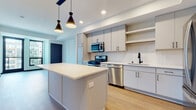 This thoughtfully designed single-floor residence showcases a clean, contemporary aesthetic throughout. The heart of the space is a well-appointed open-concept kitchen, anchored by a central island with a crisp white quartz countertop and stylish pendant lighting overhead. Light gray cabinetry lines the walls, complemented by open shelving, a textured white tile backsplash, and a full suite of stainless steel appliances including a gas range, built-in microwave, dishwasher, and refrigerator. A matte black faucet adds a refined contrast at the undermount sink. The kitchen flows seamlessly into the living area, which is bathed in recessed lighting and finished with warm-toned hardwood flooring that extends throughout the main areas. The bedroom offers a quieter retreat, featuring plush carpeting, a ceiling fan, and a large window framing elevated city skyline views that fill the room with natural light. The bathroom is finished with a clean white palette, featuring a tub-shower combination with white tile surround, a floating vanity with white cabinetry, and striking matte black hardware and fixtures that provide bold contrast. Together, the spaces deliver a cohesive, modern living environment with high-quality finishes and a functional layout.