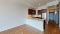 This single-floor residence showcases a clean, contemporary design with a cohesive palette of neutral tones and warm wood accents. The open-concept kitchen serves as a focal point, outfitted with rich dark-walnut cabinetry extending floor to ceiling, a light quartz peninsula with an undermount sink, a tiled backsplash, and a full suite of stainless steel appliances including a gas range, built-in microwave, and refrigerator. The layout flows naturally into the main living area, where light hardwood-style flooring and expansive floor-to-ceiling windows invite abundant natural light and sweeping city views.

A hallway lined with wall sconce lighting connects the living spaces to the bedrooms, one of which features plush carpeting and a spacious, minimalist layout. The unit includes two bathrooms: a full bath with a soaking tub, tiled surround, and dark vanity with a white countertop, and a second bathroom equipped with a wall-mounted storage cabinet, grab bar, and matching vanity finishes. The electrical panel is conveniently accessible near the entry. Throughout, the design balances functionality with modern aesthetics, creating a cohesive and well-appointed living environment.