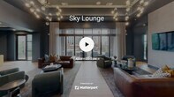 The Sky Lounge in Durham, NC is a sophisticated, single-floor communal retreat that seamlessly blends interior comfort with outdoor living. The interior is defined by rich blue-toned walls, wood-look tile flooring, and an open layout that connects the living and dining areas. The main lounge area is anchored by an arrangement of leather and upholstered seating, layered area rugs, and striking large-format abstract artwork that lends a curated, gallery-like quality to the space. Sculptural globe chandeliers overhead add a warm, ambient glow. A dedicated media room offers a more intimate setting, featuring a tray ceiling with accent lighting, a built-in diamond-pattern bookshelf with integrated greenery panels, and a wall-mounted television — all framed by deep teal walls and natural light from a large window. Throughout the interior, a cohesive palette of slate blue, teal, and warm wood tones creates an inviting and polished atmosphere. The lounge opens directly onto a spacious outdoor terrace through full-height glass doors. The terrace, framed by exposed brick columns and glass-panel railings, is furnished with seating areas and offers sweeping elevated views of the Durham skyline and surrounding green spaces, making it an ideal setting for both relaxation and socializing.