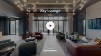The Sky Lounge in Durham, NC is a sophisticated, single-floor communal retreat that seamlessly blends interior comfort with outdoor living. The interior is defined by rich blue-toned walls, wood-look tile flooring, and an open layout that connects the living and dining areas. The main lounge area is anchored by an arrangement of leather and upholstered seating, layered area rugs, and striking large-format abstract artwork that lends a curated, gallery-like quality to the space. Sculptural globe chandeliers overhead add a warm, ambient glow. A dedicated media room offers a more intimate setting, featuring a tray ceiling with accent lighting, a built-in diamond-pattern bookshelf with integrated greenery panels, and a wall-mounted television — all framed by deep teal walls and natural light from a large window. Throughout the interior, a cohesive palette of slate blue, teal, and warm wood tones creates an inviting and polished atmosphere. The lounge opens directly onto a spacious outdoor terrace through full-height glass doors. The terrace, framed by exposed brick columns and glass-panel railings, is furnished with seating areas and offers sweeping elevated views of the Durham skyline and surrounding green spaces, making it an ideal setting for both relaxation and socializing.