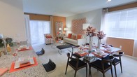 This single-floor residence offers an open-concept layout that seamlessly blends the living and dining areas into one cohesive, well-designed space. The interior is distinguished by a bold yet balanced color palette, pairing warm orange accents with neutral tones of cream and gray throughout.

The living area is furnished with a comfortable neutral sofa accented by orange throw pillows, a sleek glass-and-chrome coffee table, and a striped accent chair. A striking orange geometric wall panel serves as a focal point, adding texture and visual depth to the space. The room is bathed in natural light courtesy of large sliding glass doors dressed with floor-to-ceiling vertical blinds, which also provide access to an outdoor area.

Adjacent to the living room, the dining area features a glass-top dining table surrounded by dark wood chairs with black cushioned seats, elegantly set with place settings that echo the orange and neutral theme. Decorative floral arrangements in metallic vases add a refined finishing touch.

The space benefits from soft neutral carpeting and smooth light-toned walls, creating a bright and airy ambiance throughout. The overall design strikes a balance between modern style and warm, inviting comfort, making it both functional and visually appealing.