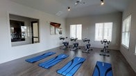 This single-floor fitness studio is thoughtfully designed to accommodate a range of exercise activities. The space features multiple stationary cycling bikes arranged in an open layout, complemented by blue exercise mats spread across the floor for stretching, yoga, or floor-based workouts. The room is finished with smooth, dark hardwood-style flooring that adds a polished, modern aesthetic while remaining practical for active use. Crisp white walls and blinds-fitted windows allow natural light to filter in, creating a bright and energizing atmosphere. A ceiling fan provides additional air circulation, ensuring comfort during intense workouts. A large mirror is mounted on one wall, a functional feature that allows participants to monitor their form during exercise. Recessed lighting and track lighting fixtures supplement the natural light, keeping the space well-lit at all times. The overall layout is clean, open, and uncluttered, maximizing usable floor space for both group and individual fitness sessions. The studio's design reflects a focus on functionality and comfort, making it a versatile environment well-suited for cycling classes, mat work, and general fitness activities.