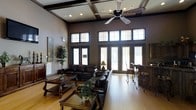 This single-story space makes a strong architectural impression with its dramatically high ceilings, detailed coffered woodwork overhead, and a ceiling fan that anchors the room's vertical scale. The main living area is generously proportioned, furnished with a suite of dark leather sofas and armchairs arranged around wooden coffee tables, creating a comfortable and cohesive seating environment. Warm-toned hardwood flooring runs throughout, complementing the dark wood trim framing the windows and doors. A bank of French doors lines the far wall, flanked by large transom windows above that flood the interior with natural light while offering views to the exterior. The tall walls are painted in a muted taupe tone, providing a neutral backdrop that enhances the richness of the darker furnishings and trim. A corner bar-height counter with wrought iron stools adds a functional social element to the space. Decorative touches include potted greenery and framed wall art, contributing to a polished yet welcoming ambiance. Overall, the space balances elegance with comfort through its architectural detailing and thoughtful furnishings.