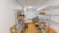 This single-floor package room is purpose-built for the secure receipt and organization of parcels and deliveries. The space is defined by its clean, utilitarian design, featuring white walls and a drop ceiling with recessed panel lighting that provides even, functional illumination throughout. The flooring consists of warm wood-look planks, offering a practical yet polished finish underfoot.

Two rows of tall, multi-tiered chrome wire shelving units run along either side of the room, creating a central aisle for easy navigation and access. These shelves accommodate a variety of package sizes, from small envelopes to large boxes, and allow for clear visibility of stored items. Additional packages are stacked along the back wall, demonstrating the room's capacity to handle substantial delivery volumes.

Entry to the room is controlled by a wall-mounted Luxer One smart package locker access panel affixed beside a solid door with a lever-style handle, enabling secure and trackable access. A small security camera is mounted near the ceiling, further reinforcing the room's focus on safety and accountability. The overall layout is straightforward and efficient, designed to maximize storage capacity while maintaining an organized and accessible environment for package management.