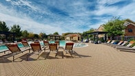 Located at 1550 Katy Gap Rd in Katy, TX, this single-story outdoor amenity complex delivers a resort-caliber experience designed for relaxation and recreation. At the heart of the space is a generously sized, freeform swimming pool with crystal-clear blue water, complemented by a separate spa with water features. The surrounding pool deck is finished in decorative paver stonework and lined with an abundance of lounge chairs and chaise lounges, providing ample space for sunbathing and leisure. Multiple pergola structures are positioned around the pool area, offering shaded retreats with seating for smaller gatherings. The clubhouse building anchors the complex, showcasing a traditional architectural style with a pitched, shingle roof, cream stucco and stone exterior, and expansive floor-to-ceiling glass walls that allow natural light to flood the interior. Decorative blue ceramic planter vessels accent the entrance courtyard, adding a touch of color to the neutral palette. The fully fenced pool area is framed by lush mature trees and tropical palms, providing both privacy and a vibrant natural backdrop. Residential buildings are visible in the surrounding area, reinforcing the community-centered character of this well-appointed outdoor amenity space.