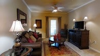 Located at 15 Brasseler Blvd in Savannah, Georgia, this single-floor residence offers a welcoming and well-appointed living environment with a classic, traditional aesthetic. The living area serves as the heart of the space, showcasing warm golden-toned walls accented by crisp crown molding that adds a refined architectural touch. A ceiling fan with a light fixture provides both comfort and ambient illumination, complementing the natural light that filters in through a glass-paned door framed by elegant floor-length drapes. Rich, dark wood furnishings — including a substantial media console and a rounded coffee table — lend a sense of warmth and sophistication. Plush leather sofas dressed with decorative pillows invite relaxation, while a patterned area rug anchors the seating arrangement and adds a layer of color and texture to the space. Tasteful wall art and accent lamps contribute to the cozy, curated ambiance throughout. The overall design reflects a cohesive traditional style, blending comfort with classic elegance. The layout is thoughtfully arranged to maximize functionality while maintaining an inviting atmosphere suited to everyday living in one of Savannah's established communities.