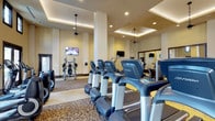 The fitness center at Lunda Vista in San Diego is a well-appointed exercise facility designed to support a comprehensive workout experience. The space features a refined interior with coffered ceilings accented by recessed and pendant lighting, creating a bright and welcoming atmosphere. Warm, neutral tones on the walls and richly patterned carpeting contribute to an upscale aesthetic that sets this gym apart from a typical fitness facility.

A row of modern cardio machines, including treadmills equipped with individual display consoles, lines the main floor and serves as the centerpiece of the space. Strength and resistance training machines are positioned throughout, providing a well-rounded selection of equipment for users of all fitness levels. Full-length mirrors along one wall visually expand the space and allow members to monitor their form during workouts.

Multiple wall-mounted flat-screen televisions are strategically placed throughout the room, ensuring visibility from nearly any position in the facility. The layout is thoughtfully organized to allow comfortable movement between equipment without crowding. Structural columns are integrated seamlessly into the design, maintaining an open and airy feel. Overall, the fitness center combines functional equipment with an elegant environment, offering residents a premium amenity for their daily wellness routines.