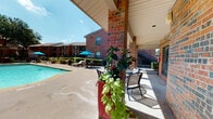 This inviting outdoor community space centers around a generously sized swimming pool set within a well-maintained courtyard. The pool area is surrounded by a broad concrete deck that provides ample room for lounging and socializing. Multiple seating arrangements are available throughout the space, including cushioned lounge chairs, shaded umbrella tables in signature blue, and a covered patio area supported by sturdy brick columns. The covered patio offers a comfortable retreat from the sun, furnished with bistro-style tables and chairs, and accented by vibrant potted plants that add a lively touch of greenery to the space. The surrounding landscape features neatly trimmed hedges, mature trees, and decorative planters with lush trailing foliage, contributing to a clean and welcoming atmosphere. The courtyard is enclosed by two-story brick residential buildings with classic architectural detailing, including pitched rooflines, exterior staircases, and balconies. A community building with large windows and a glass entry door is accessible directly from the pool deck and features posted community safety information. The entire area benefits from abundant natural sunlight and open sky views, creating a bright, resort-like ambiance ideal for outdoor recreation and relaxation.