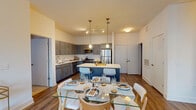 Located at 112 Southfield Ave in Stamford, Connecticut, this single-floor, two-bedroom, two-bathroom residence offers a well-designed layout with modern finishes throughout. The open-concept kitchen and dining area serves as the heart of the home, featuring gray shaker-style cabinetry, a white subway tile backsplash, stainless steel appliances including a gas range, microwave, and refrigerator, and a central island with quartz countertops and bar seating. Pendant lighting and track lighting illuminate the space, which flows seamlessly into a dedicated dining area and an adjacent living room. The living room is bright and inviting, benefiting from large windows that bring in natural light and offer outdoor views, complemented by hardwood-style flooring throughout the main areas. Both bedrooms are comfortably sized, featuring plush carpeting and large windows. Each bathroom is finished with clean, neutral tones; one features a gray vanity with a white countertop and tile flooring, while the other includes a full-size tub and shower combination with wood-look flooring. A walk-in pantry or storage closet is also visible off the kitchen, adding practical functionality to this well-appointed Stamford residence.