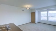 This single-floor space presents a clean, minimalist interior characterized by its neutral color palette and straightforward layout. The main room features wall-to-wall carpeting in a light beige tone, contributing to a warm and understated atmosphere throughout. Crisp white walls are complemented by white baseboards that frame the room cleanly along the floor line.

A centrally positioned pendant light fixture with a dark metal frame and bowl-shaped shade serves as the primary overhead lighting source, casting even illumination across the space. A textured ceiling adds subtle visual interest above.

A natural wood-toned door provides access to an adjoining area, offering a warm contrast against the white walls. A ceiling-mounted HVAC vent is visible, indicating a central air system integrated into the architecture.

On the far side of the room, a window allows natural light to enter, brightening the space and offering a view to the exterior. A radiator or heating unit is positioned beneath the window, suggesting supplemental heating capability. The overall ambiance is neutral and functional, making the space highly adaptable to a variety of uses and furnishing styles.
