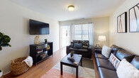 Located at 1920 Exchange Dr in Greenville, NC, this single-floor unit offers a well-designed, functional layout across multiple rooms. The living area serves as the heart of the home, featuring warm hardwood-style flooring, a wall-mounted flat-screen TV, and a sliding glass door that floods the space with natural light and provides access to an outdoor area. The open-concept kitchen is equipped with stainless steel appliances — including a refrigerator, electric range, and microwave — complemented by warm wood cabinetry and a marble-topped peninsula with a double sink, ideal for both cooking and casual dining. Hardwood-style flooring flows seamlessly from the kitchen into the main living spaces. The unit includes multiple bedrooms, each with ceiling fans and natural light from windows with blinds and sheer curtains. One bedroom features a built-in closet with wire shelving for ample storage. Two bathrooms are finished with tile flooring, wood cabinetry, and shower-tub combinations. The overall aesthetic is clean and neutral, with consistent warm tones throughout, creating a comfortable and practical living environment.