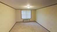 This single-floor space presents a straightforward and functional layout. The room features wall-to-wall carpeting in a neutral beige tone, providing a soft underfoot surface throughout. The walls are finished in a light cream color, accented by dark wood-toned crown molding and matching baseboards that add a subtle architectural detail along the perimeter. A textured popcorn ceiling adds dimension overhead and houses a simple flush-mount light fixture that provides central illumination to the room. A single window positioned along one wall is dressed with vertical blinds, allowing for adjustable natural light and privacy. Beneath the window sits a wall-mounted HVAC unit, offering both heating and cooling directly within the room. The overall ambiance is modest and neutral, presenting a clean, move-in-ready canvas well-suited for a variety of furnishing arrangements and uses.