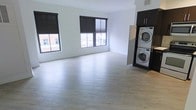 This single-floor space offers a clean, modern aesthetic defined by its open layout and minimalist design. The main living area is generously proportioned, featuring light-toned hardwood-style flooring that extends throughout, lending warmth and continuity to the space. Crisp white walls enhance the sense of openness and provide a neutral backdrop adaptable to a variety of interior styles.

Natural light is a standout feature, with multiple large windows framed in dark trim lining the exterior wall. These windows offer views of the surrounding urban streetscape and allow daylight to fill the room throughout the day. Roller shades provide privacy and light control as needed.

A particularly practical highlight is the dedicated in-unit laundry area, where a stacked washer and dryer unit is neatly integrated into a dark cabinetry enclosure. This built-in solution maximizes floor space while keeping laundry functions discreet and accessible. The dark cabinetry provides a bold contrast to the otherwise light and airy palette of the space.

Overall, the space presents a contemporary urban character, combining functional design with a bright, uncluttered ambiance well-suited for modern living.