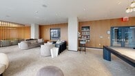 Located at 55 Clinton Pl in New Rochelle, NY, this sophisticated amenity space spans a single generously sized floor and is divided into three distinct areas, each thoughtfully designed for comfort and socializing. The café and bar area makes an immediate impression with its bold navy-blue paneled walls, channeled leather booth seating, and geometric diamond-patterned tile flooring. A curved wooden bar with a dark countertop and gold-accented pendant lighting anchors the space, while marble-topped tables and floor-to-ceiling windows flood the room with natural light and urban views. Intimate corner booths with wainscoting add a refined, classic touch. The lounge area transitions into a bright, open gathering space featuring floor-to-ceiling curtain wall glazing that frames sweeping outdoor views. Plush seating arrangements — including barrel chairs and a full sofa grouping — are complemented by a billiards table beneath a striking cluster chandelier with gold detailing. A media lounge rounds out the space, showcasing curved warm-wood wall paneling, a large flat-screen display, and a sculptural green curved sofa paired with a travertine coffee table, creating an inviting, cocoon-like atmosphere. Throughout, the design balances contemporary elegance with relaxed, residential warmth.