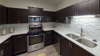 This single-floor residence offers a thoughtfully designed layout that balances functionality and modern style. The open-concept living area is finished with plush neutral carpeting and painted in soft, warm tones, creating a welcoming atmosphere. Track lighting overhead adds a contemporary touch, while the elevated ceiling detail near the kitchen transition adds visual interest and a sense of added height.

The kitchen is a standout feature, equipped with dark espresso cabinetry accented by sleek hardware, light granite countertops, and a textured tile backsplash. Stainless steel appliances — including a gas range, over-the-range microwave, and dishwasher — complete the space. Wood-look flooring grounds the kitchen and extends into the connecting hallway, providing a durable and attractive surface.

The hallway connects to a well-organized bathroom corridor featuring granite countertops, dark cabinetry, and wood-look tile flooring, with multiple doorways providing practical access to adjacent rooms. The bedroom is generously sized, carpeted, and benefits from a large window that invites natural light and offers pleasant greenery views. A ceiling fan adds comfort and utility to the space.