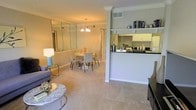 This single-floor residence showcases a thoughtfully designed open-concept layout that seamlessly connects the living, dining, and kitchen areas into one cohesive and functional living space. The living area features plush carpeting in a soft neutral tone, providing a warm foundation for the room, and is complemented by elegant crown molding that frames the ceilings throughout. A marble-top coffee table and coordinating furnishings add a refined touch to the space.

The dining area is anchored by a round table with sleek chairs and is illuminated by a classic chandelier that adds a decorative focal point overhead. A large floor-to-ceiling mirror wall along the dining area reflects light beautifully, enhancing the sense of openness and brightness throughout the space.

The kitchen is positioned adjacent to the dining room and features a half-wall pass-through with a dark countertop, cream-toned cabinetry, and open display shelving above — ideal for both practical storage and decorative accents. The layout promotes easy interaction between the kitchen and living spaces, making it well-suited for entertaining.

Consistent crown molding, neutral wall tones, and coordinated finishes throughout give this residence a polished and cohesive aesthetic from every angle.