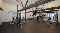 Located at 2858 W Ball Rd in Anaheim, California, this single-story fitness center offers a thoughtfully designed workout space with a striking architectural character. The room's most defining feature is its vaulted ceiling adorned with bold, dark exposed beam trusses that span the width of the space, creating an open and airy atmosphere while adding a distinct visual appeal.

The fitness center is equipped with a comprehensive selection of exercise equipment, including a multi-station cable strength machine, multiple treadmills, stationary bikes, an elliptical trainer, and additional free-weight and bench stations. The flooring throughout is durable rubber matting, ideal for a high-use fitness environment. Large mirrors line one wall, enhancing the sense of space and allowing users to monitor their form during workouts. Ceiling fans are positioned throughout, contributing to comfortable airflow during exercise.

Natural light enters through multiple windows fitted with horizontal blinds, offering views of the surrounding greenery. A set of glass double doors at one end of the space provides direct access to an outdoor patio area, further brightening the interior. Recessed lighting and white paneled walls complete the clean, functional aesthetic of this well-appointed facility.