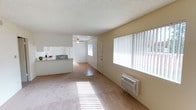 This single-floor space offers an open, airy layout that seamlessly connects the living area, dining space, and kitchen. The living area is finished with plush carpeting and is bathed in natural light streaming through a large window dressed with vertical blinds. A wall-mounted air conditioning unit provides climate control convenience. The flooring transitions from carpet in the living zone to hardwood-style flooring as you move toward the kitchen and dining areas, adding visual distinction between the spaces. The open-concept kitchen features a raised breakfast bar countertop that serves as a natural divider, white cabinetry, and direct sightlines into the main living area, making the layout feel expansive and connected. A ceiling fan with a light fixture is positioned overhead in the dining area, enhancing airflow and illumination. The textured popcorn ceiling runs throughout, and the neutral, cream-toned walls create a clean, versatile backdrop. A paneled door near the rear of the space provides access to an additional room or exit. Overall, the space presents a functional, well-lit interior with a straightforward, open floor plan.