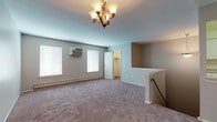 This single-floor unit offers a well-organized layout comprising a living area, kitchen, multiple bedrooms, and a full bathroom. The living area is highlighted by a chandelier overhead and plush wall-to-wall carpeting underfoot, lending a warm, comfortable feel to the space. A staircase railing detail adds a subtle architectural touch, and windows with blinds allow natural light to filter in while baseboard heating and wall-mounted air conditioning units ensure year-round climate control.

The kitchen is efficiently designed with ample white cabinetry providing generous storage above and below the countertops. A coil-burner electric range, a black dishwasher, and a single-basin sink with a standard faucet complete the functional setup. Tile flooring adds durability to this work area.

The bedrooms are carpeted and feature neutral-toned walls, creating a calm, versatile environment. One bedroom benefits from natural light through two windows and is equipped with a wall A/C unit, while double bi-fold closet doors offer substantial storage. A second bedroom also features carpeting and a doorway leading to an adjacent hallway. The full bathroom is finished cleanly in white, featuring a tub/shower combination, a single-sink vanity with white cabinetry, and tile flooring.