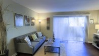 This single-story space centers around a well-appointed living room that balances comfort and contemporary style. The room is anchored by a spacious gray sofa dressed with an assortment of decorative throw pillows in complementary patterns and colors, positioned across from a rustic wood-framed coffee table resting atop a plush area rug. A tall floor lamp provides warm ambient lighting, supplementing the abundant natural light that filters through large vertical-blind-covered sliding glass doors along one wall. These expansive doors serve as a prominent architectural feature, drawing in daylight and offering potential access to an outdoor area. The walls are adorned with a curated selection of framed artwork and canvas panels, adding visual interest and personality to the neutral, softly painted walls. The overall ambiance of the space is welcoming and relaxed, with a cohesive color palette of cool grays and muted greens that contributes to a calm, livable atmosphere. The open layout and thoughtful furnishings make efficient use of the available floor space, creating a functional yet aesthetically pleasing environment suitable for everyday living.