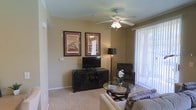 This single-floor space features a well-appointed living room that serves as the central gathering area. The room is finished with wall-to-wall carpeting in a neutral tone, contributing to a warm and comfortable atmosphere. A ceiling fan with an integrated light fixture is centrally positioned, providing both air circulation and ambient lighting. Warm beige walls create a welcoming backdrop throughout the space.

The living room is thoughtfully arranged with a dark-finished media console housing a flat-screen television, complemented by two framed art pieces mounted above. A floor lamp adds additional ambient lighting, while a glass-top coffee table and upholstered seating, including a sofa and accent chair, complete the furnishings. Vertical blinds dress a large sliding glass door that allows natural light to filter into the room and provides access to an outdoor area, offering views of the surrounding landscape. The combination of neutral finishes, soft carpeting, and natural light creates a relaxed, functional living environment.