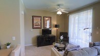 This single-floor space features a well-appointed living room that serves as the central gathering area. The room is finished with wall-to-wall carpeting in a neutral tone, contributing to a warm and comfortable atmosphere. A ceiling fan with an integrated light fixture is centrally positioned, providing both air circulation and ambient lighting. Warm beige walls create a welcoming backdrop throughout the space.

The living room is thoughtfully arranged with a dark-finished media console housing a flat-screen television, complemented by two framed art pieces mounted above. A floor lamp adds additional ambient lighting, while a glass-top coffee table and upholstered seating, including a sofa and accent chair, complete the furnishings. Vertical blinds dress a large sliding glass door that allows natural light to filter into the room and provides access to an outdoor area, offering views of the surrounding landscape. The combination of neutral finishes, soft carpeting, and natural light creates a relaxed, functional living environment.