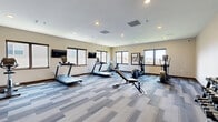Located at 2109 E Anamosa St in Rapid City, South Dakota, this fitness center offers a dedicated, single-floor exercise space designed to support a comprehensive workout routine. The room is finished with a modern patterned carpet in neutral blue and gray tones, complemented by warm beige walls and dark wood baseboards that lend a clean, contemporary feel throughout.

Natural light fills the space through multiple large windows positioned along the perimeter walls, offering views of the surrounding Rapid City landscape and creating an open, energizing atmosphere. Recessed ceiling lighting supplements natural illumination, ensuring the space remains bright at all hours.

The exercise room is well-appointed with a variety of cardiovascular equipment, including multiple treadmills, an elliptical trainer, and a stationary bike. A weight bench and a dumbbell rack with a full set of free weights provide strength training options. Wall-mounted flat-screen televisions are positioned for convenient viewing during cardio sessions. Motivational wall art and whiteboards add a purposeful, focused ambiance to the room.

The layout is thoughtfully organized to maximize usable floor space, allowing comfortable movement between equipment. A dedicated entry door, ventilation systems, and a sprinkler system reflect attention to safety and comfort. Overall, this fitness center presents a functional and inviting environment for residents seeking convenient on-site exercise facilities.
