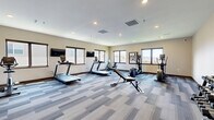 Located at 2109 E Anamosa St in Rapid City, South Dakota, this fitness center offers a dedicated, single-floor exercise space designed to support a comprehensive workout routine. The room is finished with a modern patterned carpet in neutral blue and gray tones, complemented by warm beige walls and dark wood baseboards that lend a clean, contemporary feel throughout.

Natural light fills the space through multiple large windows positioned along the perimeter walls, offering views of the surrounding Rapid City landscape and creating an open, energizing atmosphere. Recessed ceiling lighting supplements natural illumination, ensuring the space remains bright at all hours.

The exercise room is well-appointed with a variety of cardiovascular equipment, including multiple treadmills, an elliptical trainer, and a stationary bike. A weight bench and a dumbbell rack with a full set of free weights provide strength training options. Wall-mounted flat-screen televisions are positioned for convenient viewing during cardio sessions. Motivational wall art and whiteboards add a purposeful, focused ambiance to the room.

The layout is thoughtfully organized to maximize usable floor space, allowing comfortable movement between equipment. A dedicated entry door, ventilation systems, and a sprinkler system reflect attention to safety and comfort. Overall, this fitness center presents a functional and inviting environment for residents seeking convenient on-site exercise facilities.