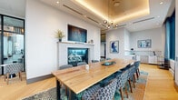 This elegantly designed single-floor space offers a refined environment ideal for gatherings, meetings, or private events. The room is anchored by a substantial natural wood conference table surrounded by stylishly patterned upholstered chairs, comfortably seating a large group. A coffered ceiling with warm ambient cove lighting adds architectural depth and a sense of luxury overhead, complemented by a contemporary multi-globe pendant chandelier.

A sleek fireplace with a light stone surround serves as a striking focal point on one wall, topped by a wall-mounted flat-screen display and flanked by tasteful decorative accents. Curated artwork adds character and sophistication throughout the space.

Floor-to-ceiling windows along the exterior wall flood the room with abundant natural light and frame sweeping panoramic city skyline views, creating a dramatic and inspiring backdrop. Sheer drapes offer flexible light control while maintaining the airy atmosphere.

The flooring features elegant light oak wood laid in a herringbone pattern, adding visual texture and warmth. A vibrant, multi-toned area rug grounds the seating area with a bold design element. A kitchenette alcove with modern built-in appliances and white cabinetry provides functional support for catered events. The overall ambiance is polished, modern, and welcoming.