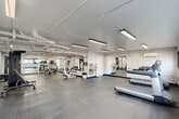 Located at 730 Germantown Cir in East Ridge, Tennessee, this fitness center offers a well-organized and fully equipped exercise room designed to support a wide range of workout routines. The space is finished with a clean, utilitarian aesthetic — white walls, exposed overhead ductwork, and bright linear fluorescent lighting that ensures consistent illumination throughout.

The durable diamond-plate rubber flooring provides secure footing across the entire floor, while full-length mirrored walls along multiple sides visually expand the room and allow users to monitor their form during exercise. Large horizontal windows along one wall invite natural light and offer views of the surrounding outdoor landscape.

The cardio zone features multiple treadmills and elliptical machines, providing ample options for aerobic training. The strength and functional training area includes a cable resistance machine, an adjustable bench, TRX suspension straps, and a variety of free weights and accessories such as stability balls. A dedicated open area offers space for stretching, floor exercises, or mat-based workouts.

Additional amenities include a water fountain, wall-mounted equipment storage, and clearly marked exit access. A motivational wall graphic reading "Stronger Yesterday" adds an encouraging touch to the overall atmosphere. The fitness center is thoughtfully arranged to accommodate multiple users simultaneously, making it a practical and versatile facility.