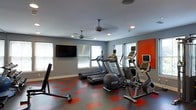 Located at 750 Summer St in Stamford, Connecticut, this fitness center offers a well-equipped and thoughtfully designed workout space on a single level. The room features a striking modern aesthetic defined by cool gray walls, a bold orange accent wall, and a dynamic gray-and-red modular tile floor that adds energy and visual interest to the space. Recessed lighting and dual ceiling fans ensure the room remains well-lit and comfortably ventilated during workouts. Large windows along the perimeter flood the space with natural light, contributing to an open and inviting atmosphere. The cardio equipment lineup includes multiple treadmills, an elliptical cross-trainer, and a stationary bike, each outfitted with individual digital display monitors for an enhanced workout experience. Free weights and additional training accessories are also available, catering to a range of fitness needs. A wall-mounted flat-screen television provides entertainment and motivation during sessions. The overall layout is clean and uncluttered, allowing for easy movement between stations. This fitness center combines functional equipment with a modern, energetic design to create a motivating environment for users of all fitness levels.